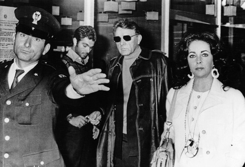 Richard Burton and Elizabeth Taylor being escorted by a policeman at Rome's Leonardo da Vinci airpport on departure for Lecce, Southern Italy 11/18/1972