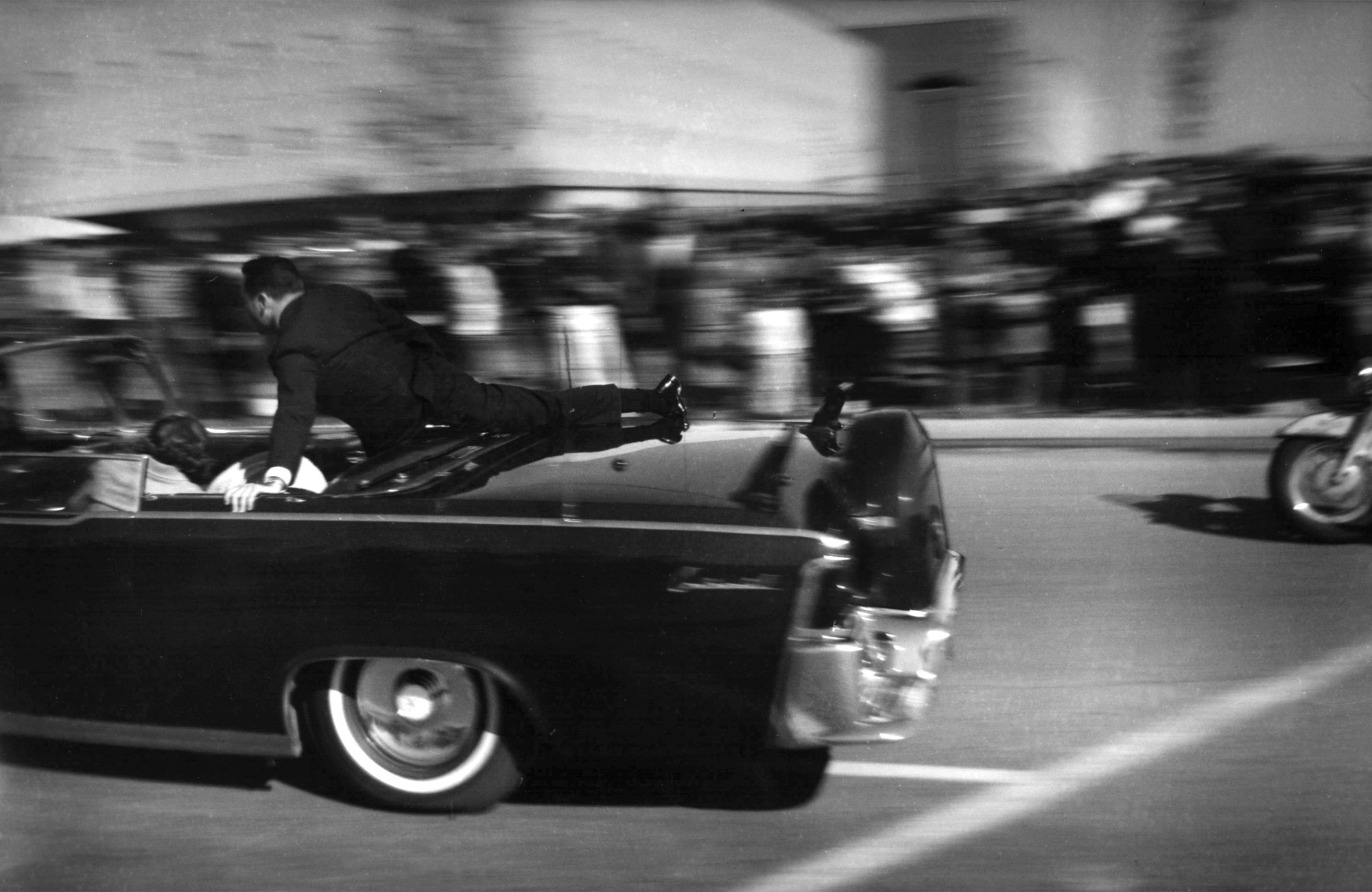Secret Service Agent Clint Hill uses his body to shield President and Mrs. Kennedy from assassin's bullets on 11/22/63 as car reaches speeds of 80 mph.