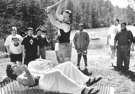 Demonstrating that his PerkiSystem total fitness program builds strong bodies, health nut Tony Perkis (Ben Stiller, supine) proves his point on a bed of nails as the awestruck campers witness Lars (Tom Hodges, center) smashing a block of ice on Tony's brick-hard stomach.