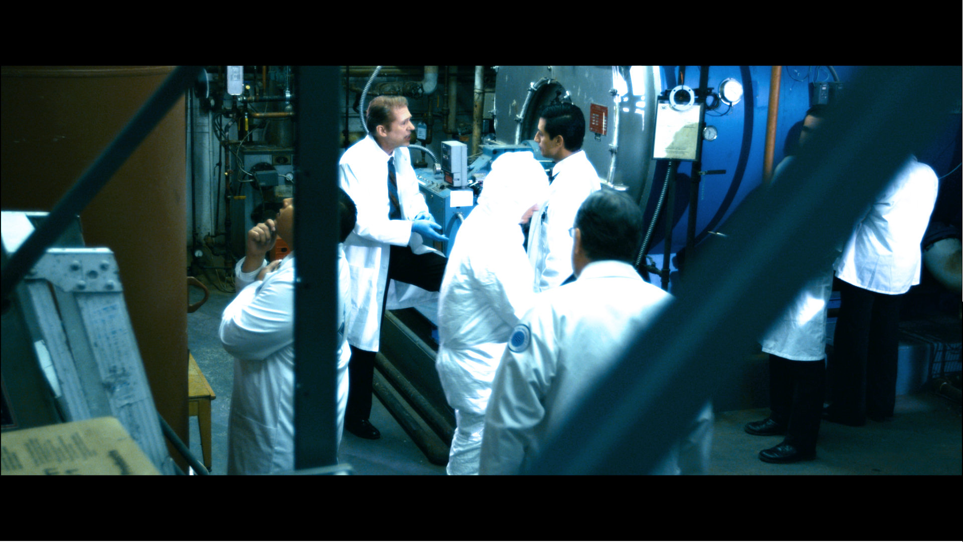 Dr. Greenglass (Allen Enlow) scolds Felix (Alex Kruz) for overlooking the little things as he holds up a filter responsible for altering the functionality of the machine. Marilyn - the nuclear reactor which Dr. Greenglass invented behind them. Several scientists and technicians in hazmat and radioactive protective gear busily whirr around their daily duties.