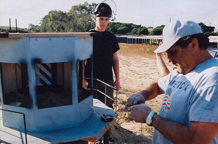 Director Gary Ambrosia looks on as pyrotechnitian John Saunders rigs a miniature for detonation.