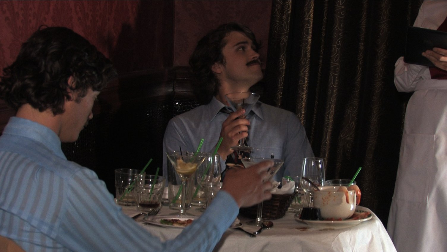 Billy (Sean Carlin), wearing a fake moustache, orders another round of amaretto and pineapple while Paul (Josiah Lipscomb) shows his fake room key to charge the drinks to his hotel room.