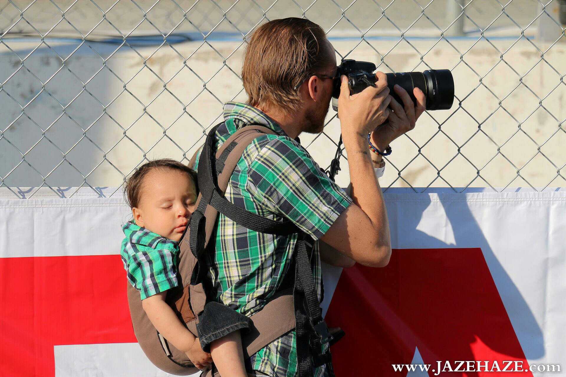 Portrait of an unknown photographer shooting races at the Toyota Grand Prix at Long Beach while his child is sound asleep in the midst of all the engine noise.