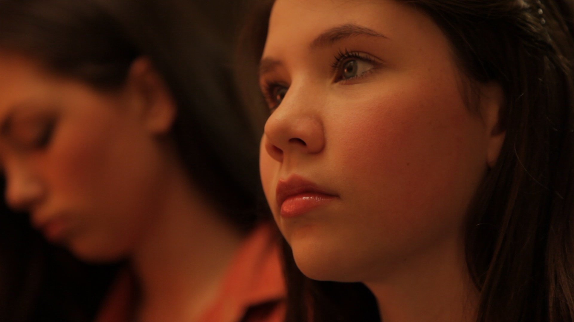 Rachel stares at the paintings on the wall during piano practice as Amy, in the background, looks through her phone.