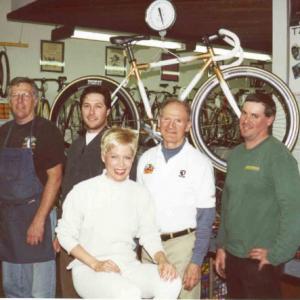At Ted Ernsts Bicycle Shop in Manhattan Beach seated in front is World Champion medallist Libby Aubrey Libby and her topnotch four man mechanic team pose with Libbys custom fully loaded carbonfiber Calfee race bike called the WHITE ANGEL hanging from the ceiling in the bike scale over head