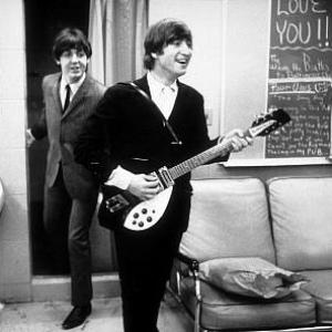 The Beatles Paul McCartney stepping out of a small room and John Lennon on the foreground playing his guitar c 1964